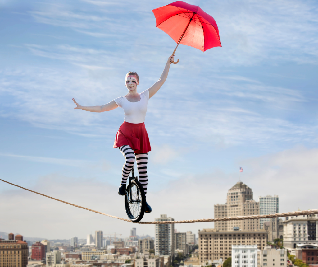 L'équilibre vie pro vie perso se manifeste souvent par un déséquilibre : d'où le choix de cette image qui montre une équilibriste sur un monocycle avec une ombrelle rouge, grimée comme une clown, essayant de se tenir en équilibre sur une corde, au-dessus d'une ville aux grands immeubles